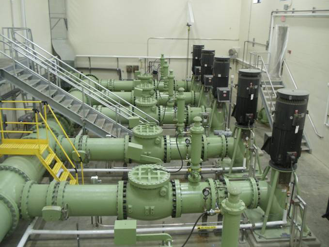 Interior view of a water pumping station with multiple parallel green pipelines running horizontally, equipped with valves, flanges, and pressure gauges. Large black electric motors are mounted vertically on the right side of each pipe assembly. Metal stairs with yellow safety handrails provide access on the left and right. The room has beige walls, exposed piping overhead, and a clean industrial floor.