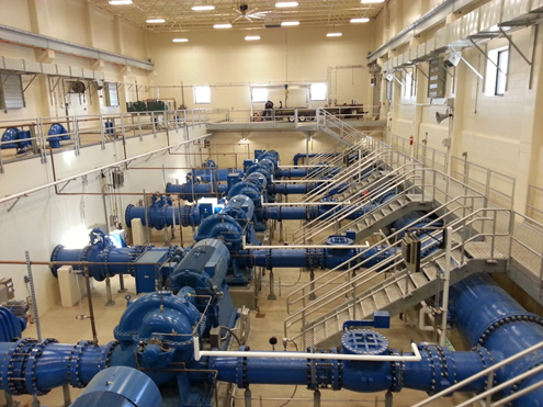 Spacious interior of a pump room featuring rows of large blue centrifugal pumps connected to horizontal blue pipelines with valves and fittings. Metal walkways and stairs with railings allow access to upper levels. Overhead fluorescent lighting illuminates the beige brick walls, high windows, and ceiling fans. A person is visible on the upper walkway in the background.